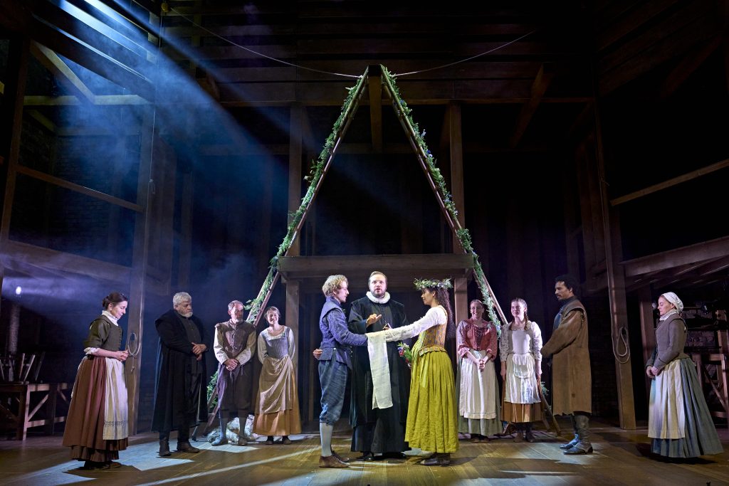 The cast of Hamnet stand on a wooden stage. Behind them, two wooden ladders fashioned into an ‘A’, draped with flowers. Photo by Manuel Harlan © RSC