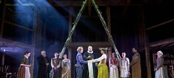 The cast of Hamnet stand on a wooden stage. Behind them, two wooden ladders fashioned into an ‘A’, draped with flowers. Photo by Manuel Harlan © RSC